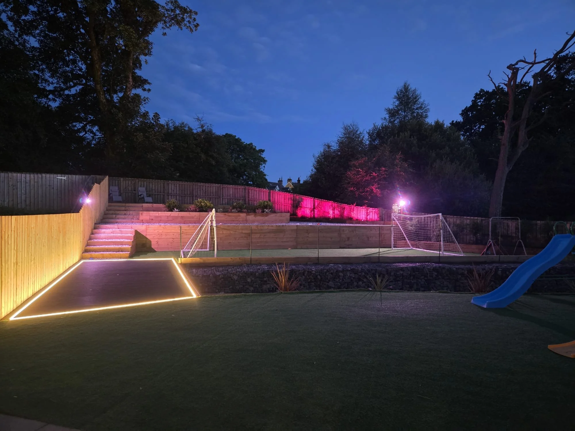 A backyard at dusk with a wooden fence, tiered garden beds, trees, and a small sports court illuminated with string lights, colorful spotlights, and a children's play slide in the corner.