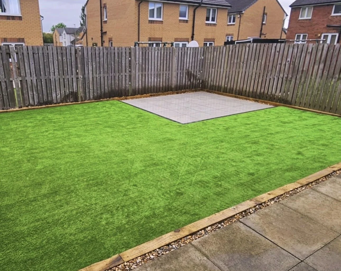 A backyard with artificial green grass, a tiled area, and a wooden fence. Surrounding houses are visible in the background.