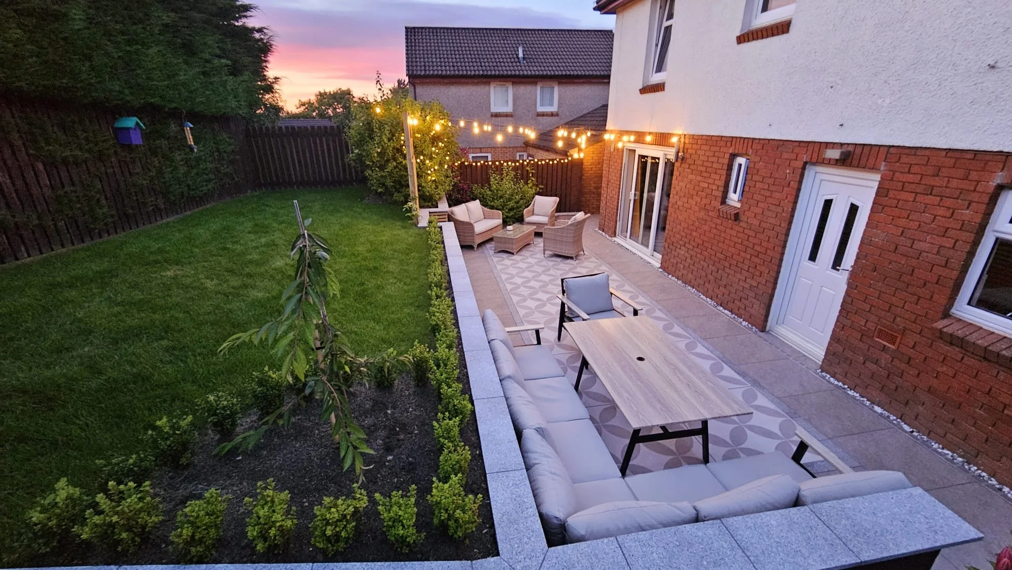A backyard patio with outdoor furniture and string lights, adjacent to a house with brick and white walls, a manicured lawn, and a sunset sky in the background.