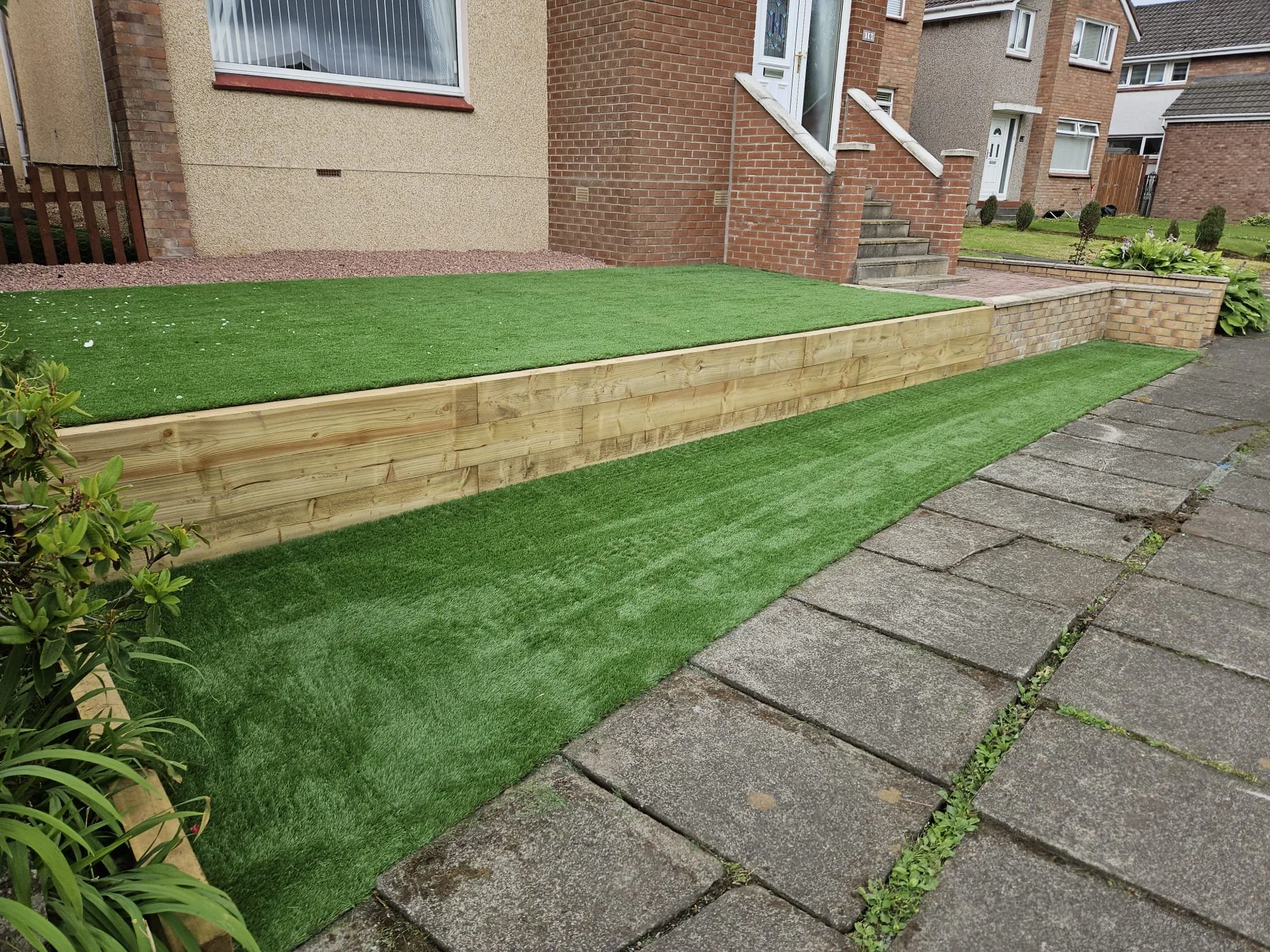 A residential front yard with artificial grass, a wooden retaining wall, stone pavers, and brick steps leading to the front door of a house.