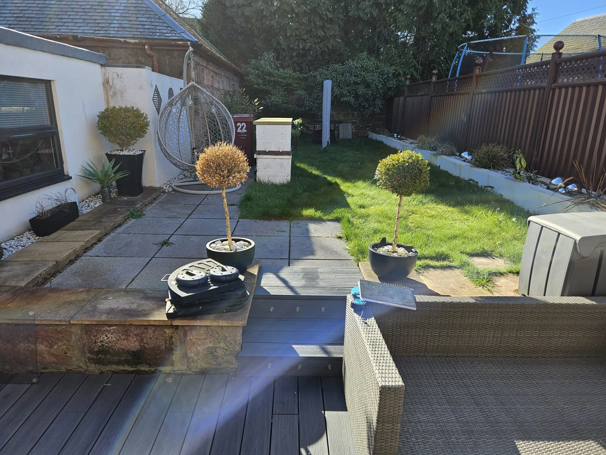 A backyard with a small lawn, potted plants, a hanging egg chair, a grill, and outdoor furniture, enclosed by a brown wooden fence and a white brick wall.