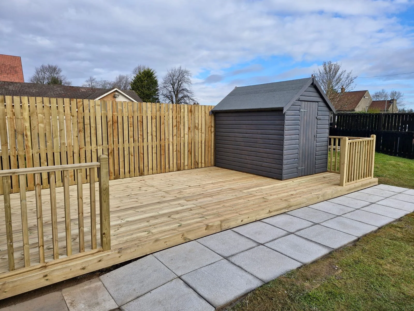 Backyard with a newly built wooden deck, a small dark gray shed, a wooden fence, and a row of concrete pavers.