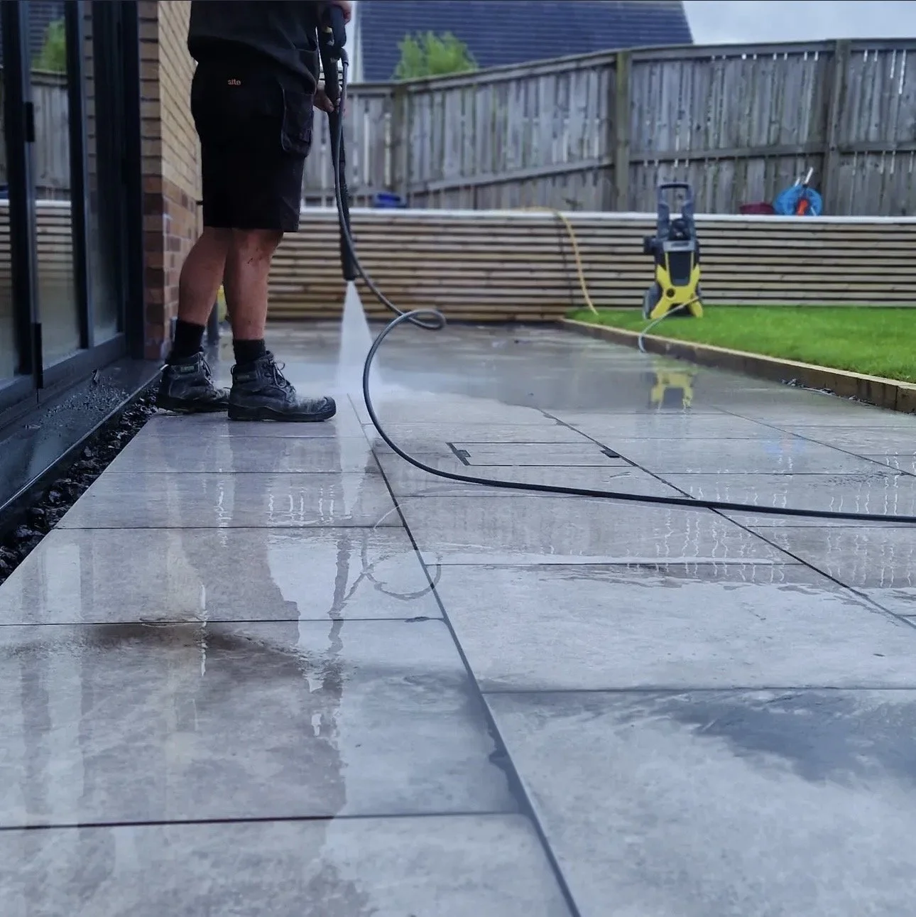 A person power washing a tiled patio in a backyard during daytime. The person is wearing black shorts, a dark shirt, black boots, and black socks, holding a power washer nozzle, with water spraying on the tiles. There is a wooden fence, some gardenin