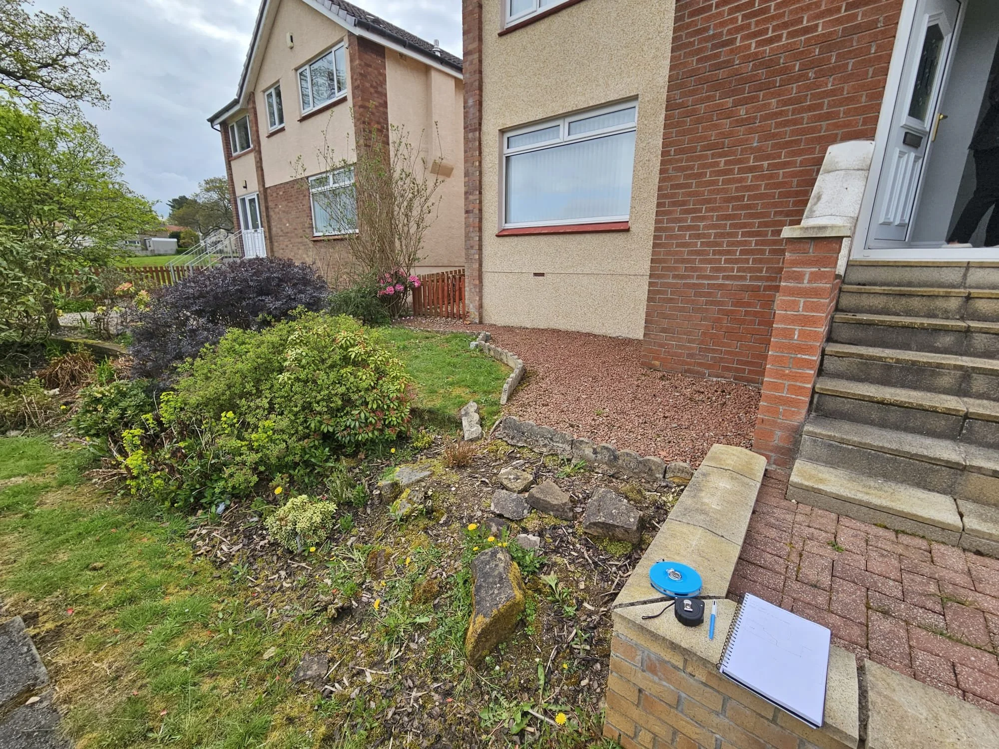 Front yard garden with various plants and shrubs, steps leading to door, some equipment and a notebook on the steps, part of a beige and brick house.