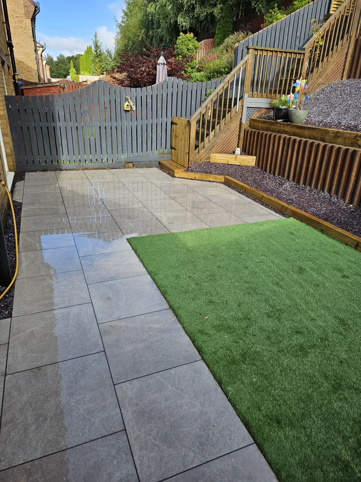 Backyard patio with gray tile flooring, green artificial grass, wooden stairs and fencing, some plants, and garden decorations under a partly cloudy sky.