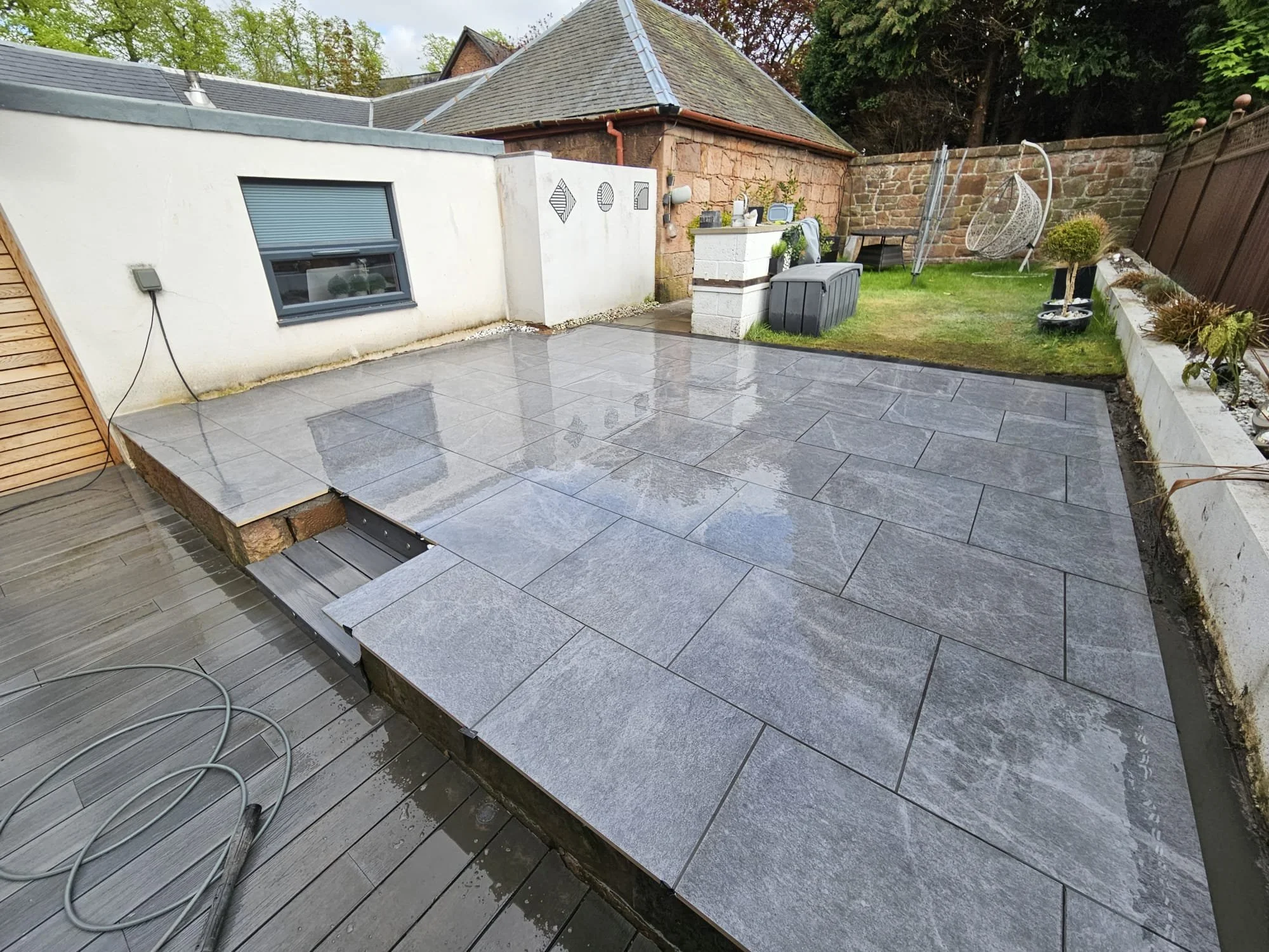 Backyard patio with new gray stone tiles, partially wet, with a wooden garden bed and a white wall with decorative black wire art pieces. There is a grassy area with a hanging chair, small trees in pots, and a brick wall with greenery behind.