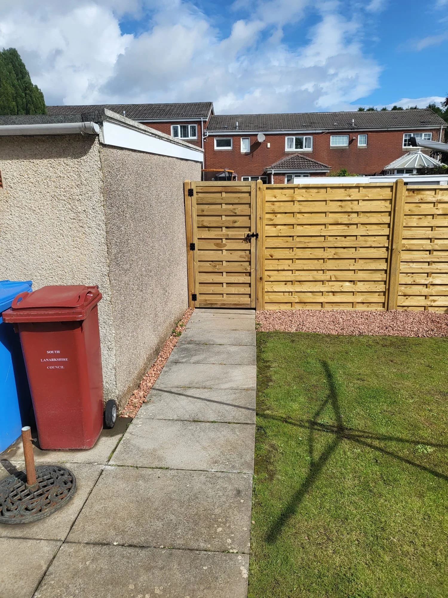 A backyard patio with a concrete walkway, a wooden gate, a red wheelie bin labeled 'South Lanarkshire Council', a portion of a textured exterior wall, and a grassy lawn on the right side. In the background, there are two-story red brick houses with tiled roofs and satellite dishes, under a partly cloudy sky.