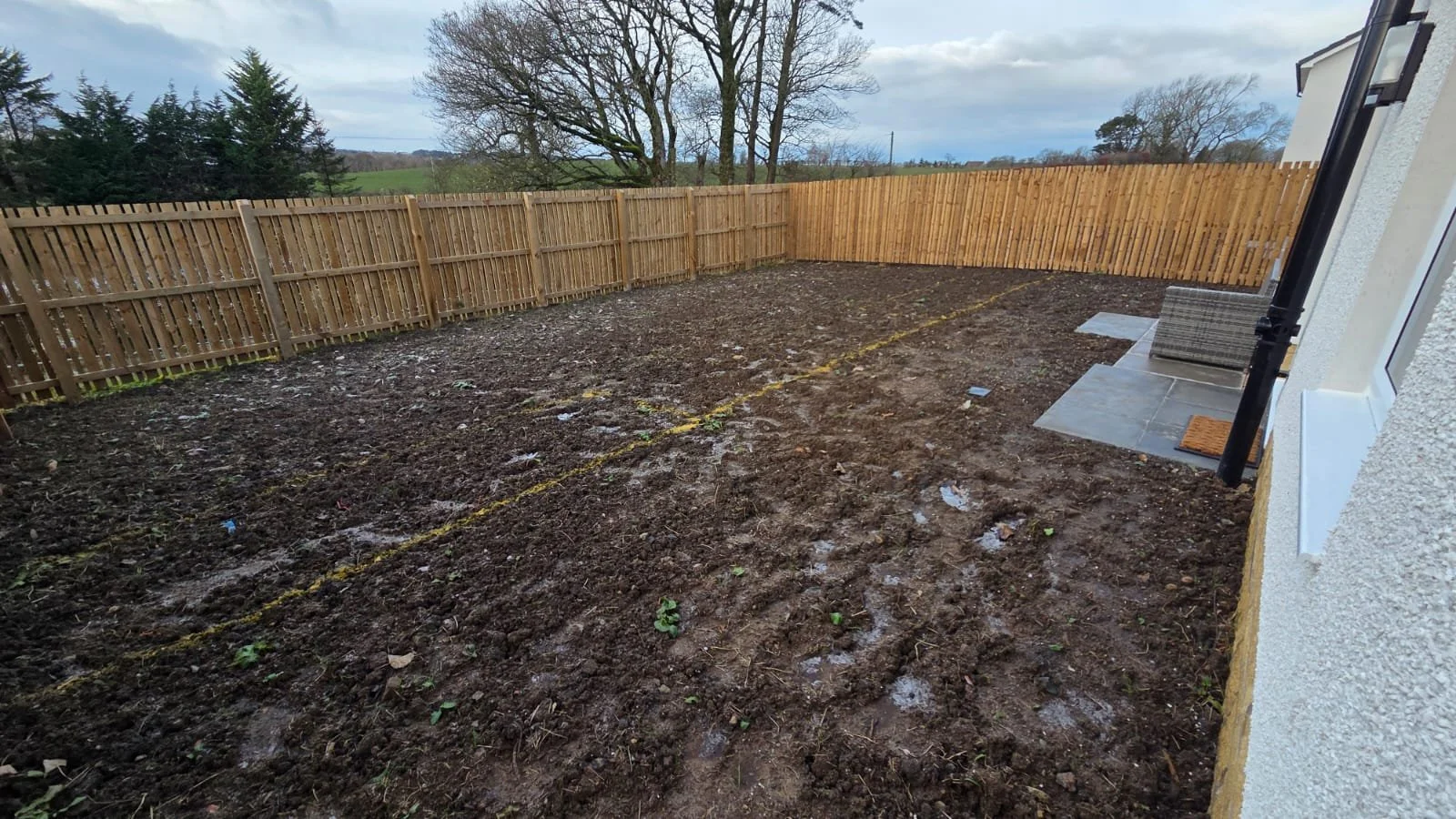 Bare backyard with freshly tilled soil, a wooden fence around the perimeter, and some small green plants starting to grow. A small patio area with outdoor furniture and a downspout on the house wall.