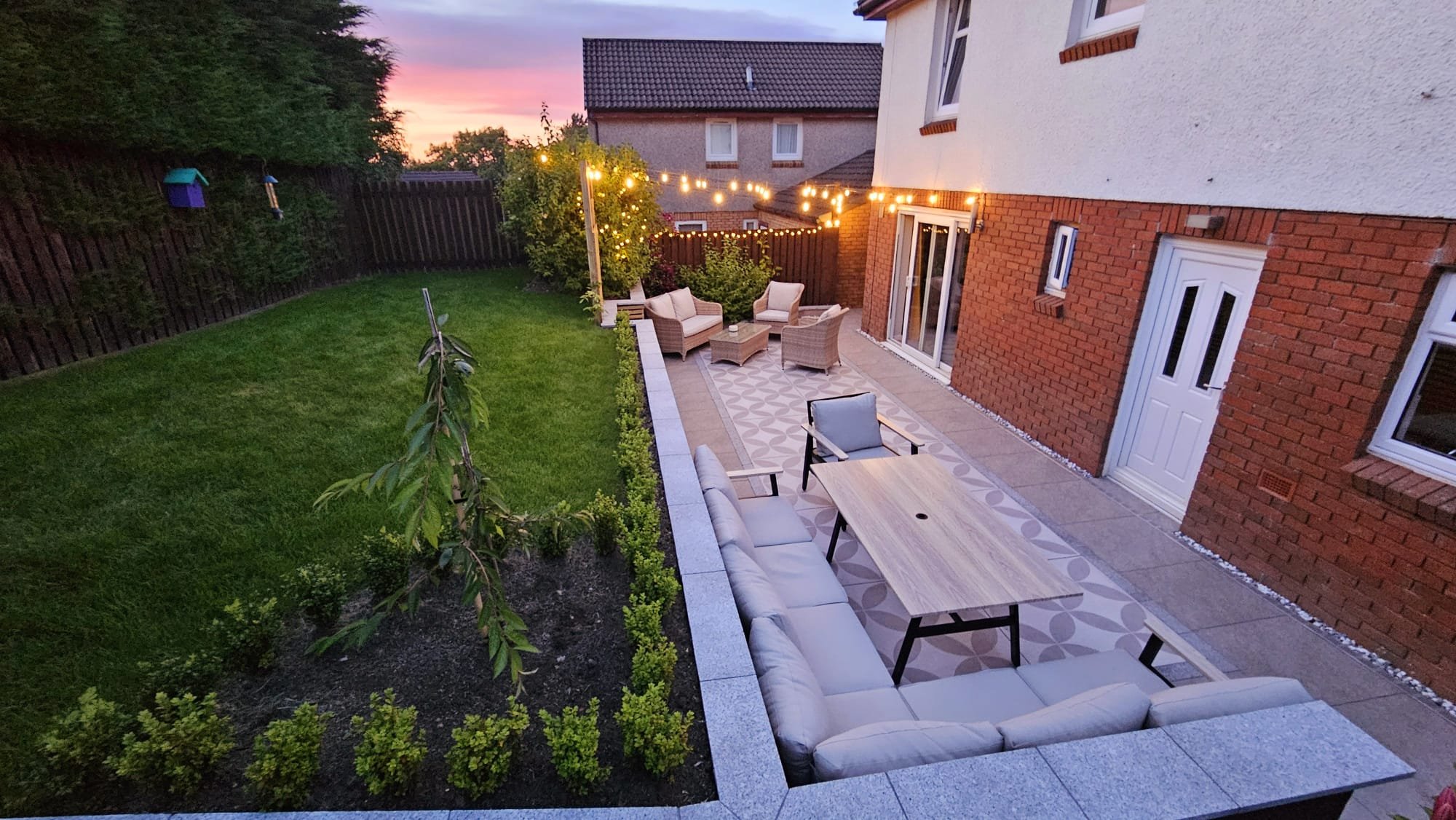 A backyard patio with outdoor seating, string lights, and a lawn, during sunset.