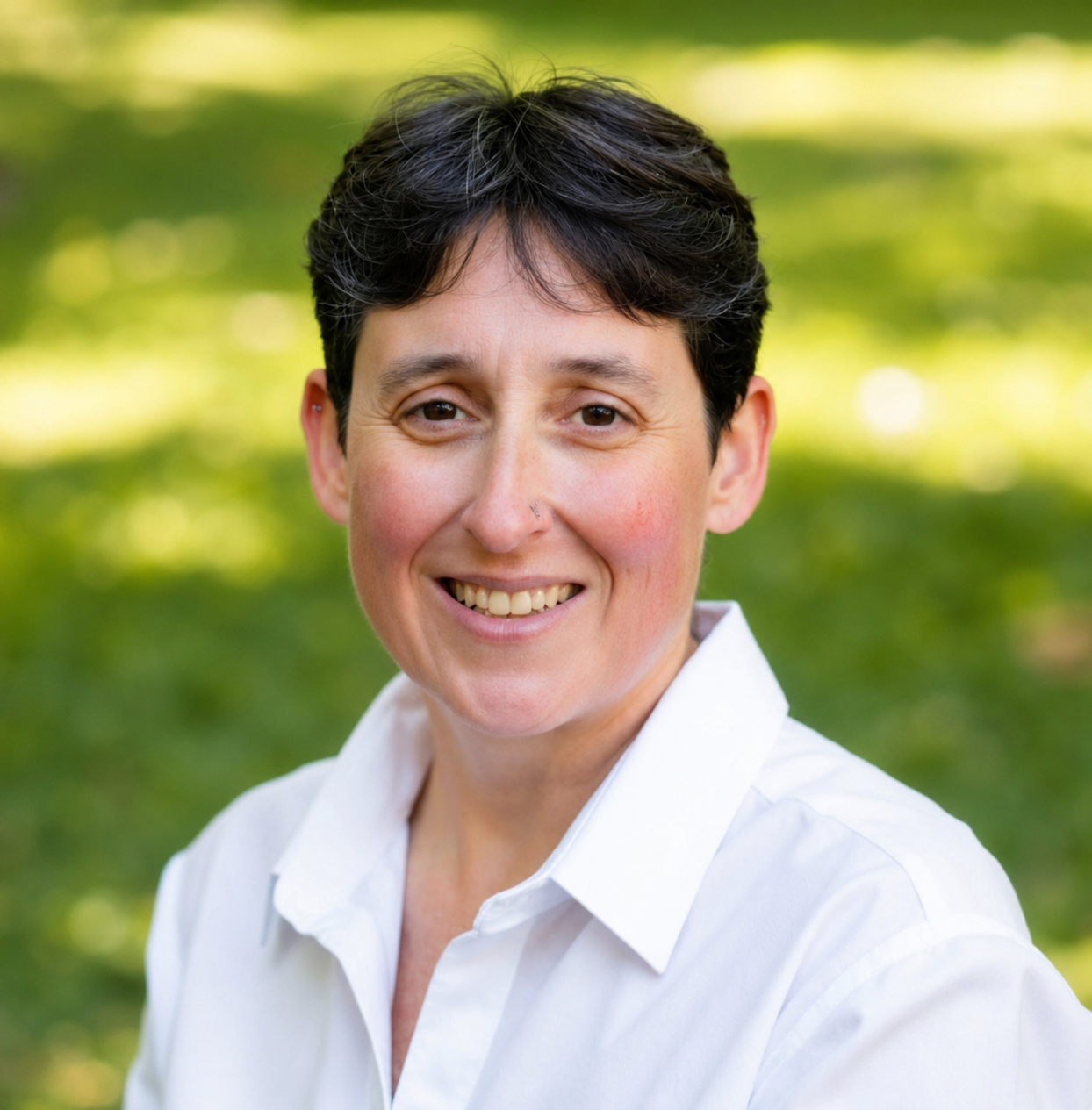 A woman with short dark hair smiling outdoors in a white collared shirt with a blurred green background.