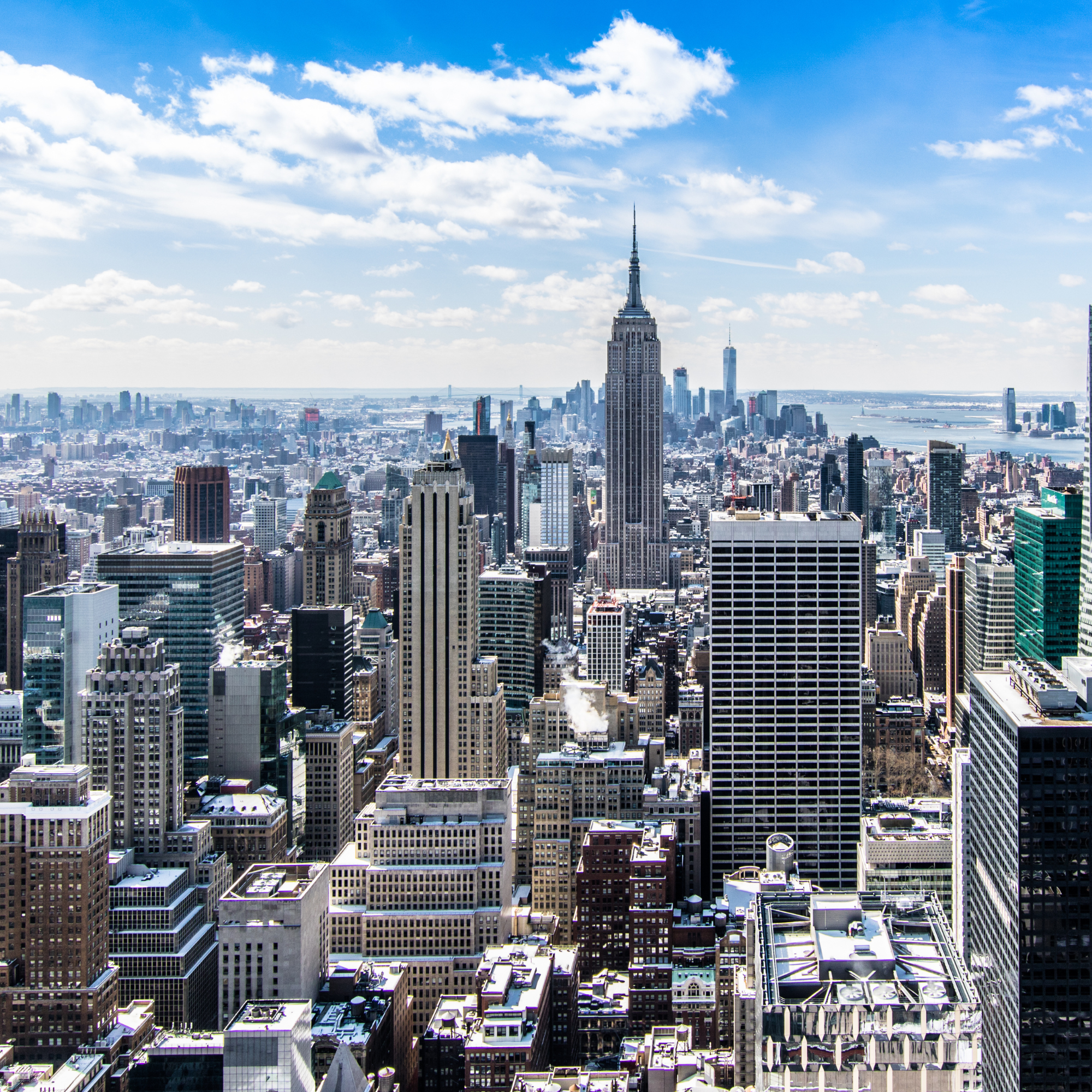 Aerial view of Manhattan skyline with the Empire State Building prominently in the center, surrounded by other tall skyscrapers and buildings, with a partly cloudy blue sky overhead.