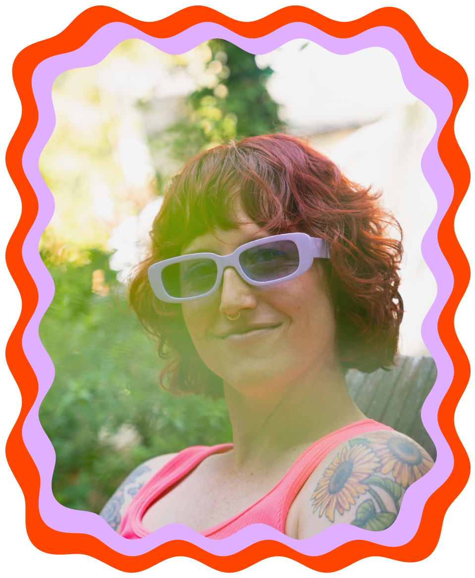 A woman with short, curly red hair wearing lavender sunglasses, a pink tank top, and colorful sunflower tattoos on her arm outdoors in sunlight.
