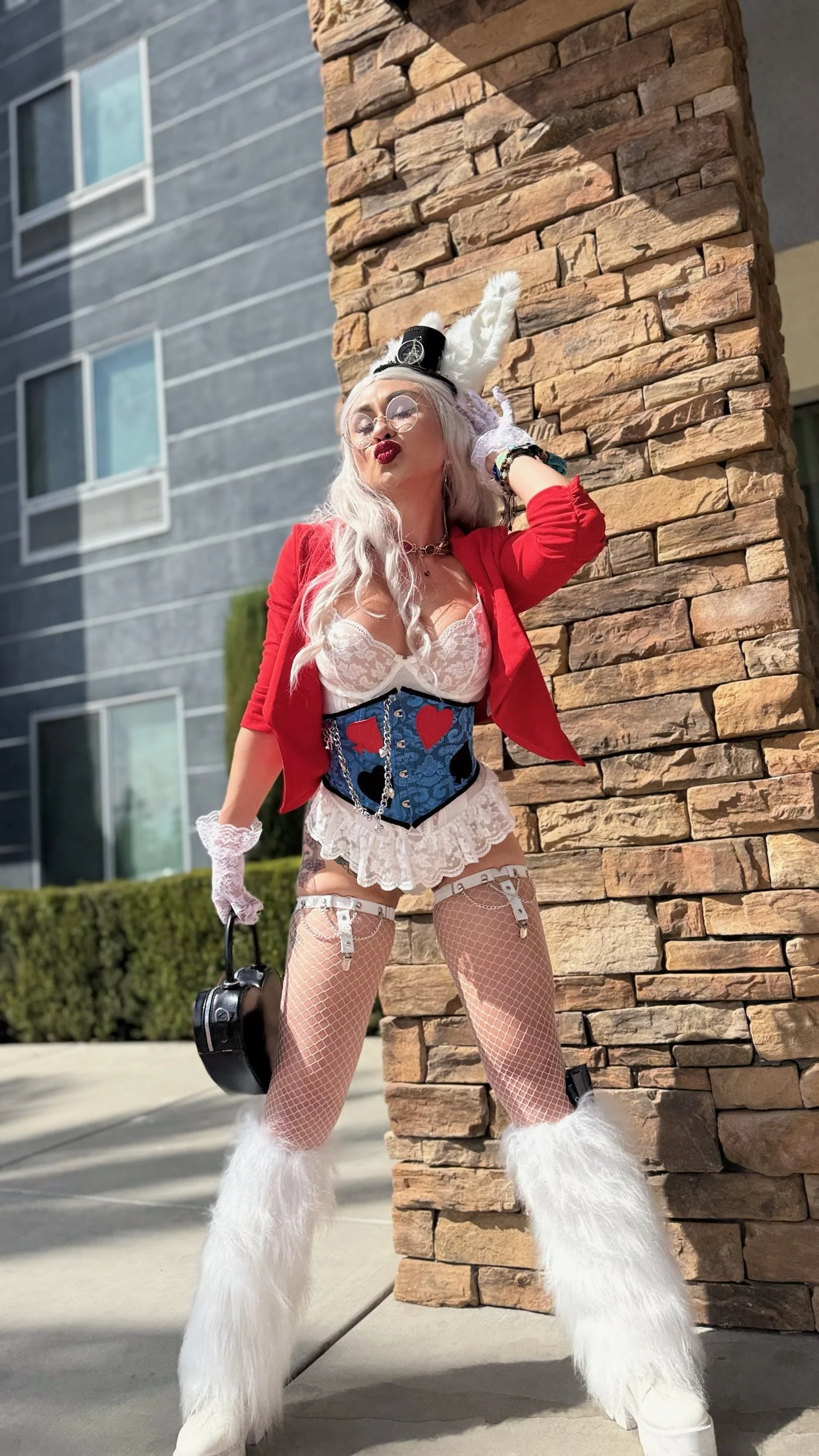 Woman dressed in a playful, costume-style outfit, standing next to a stone wall with modern building windows in the background, striking a playful pose.
