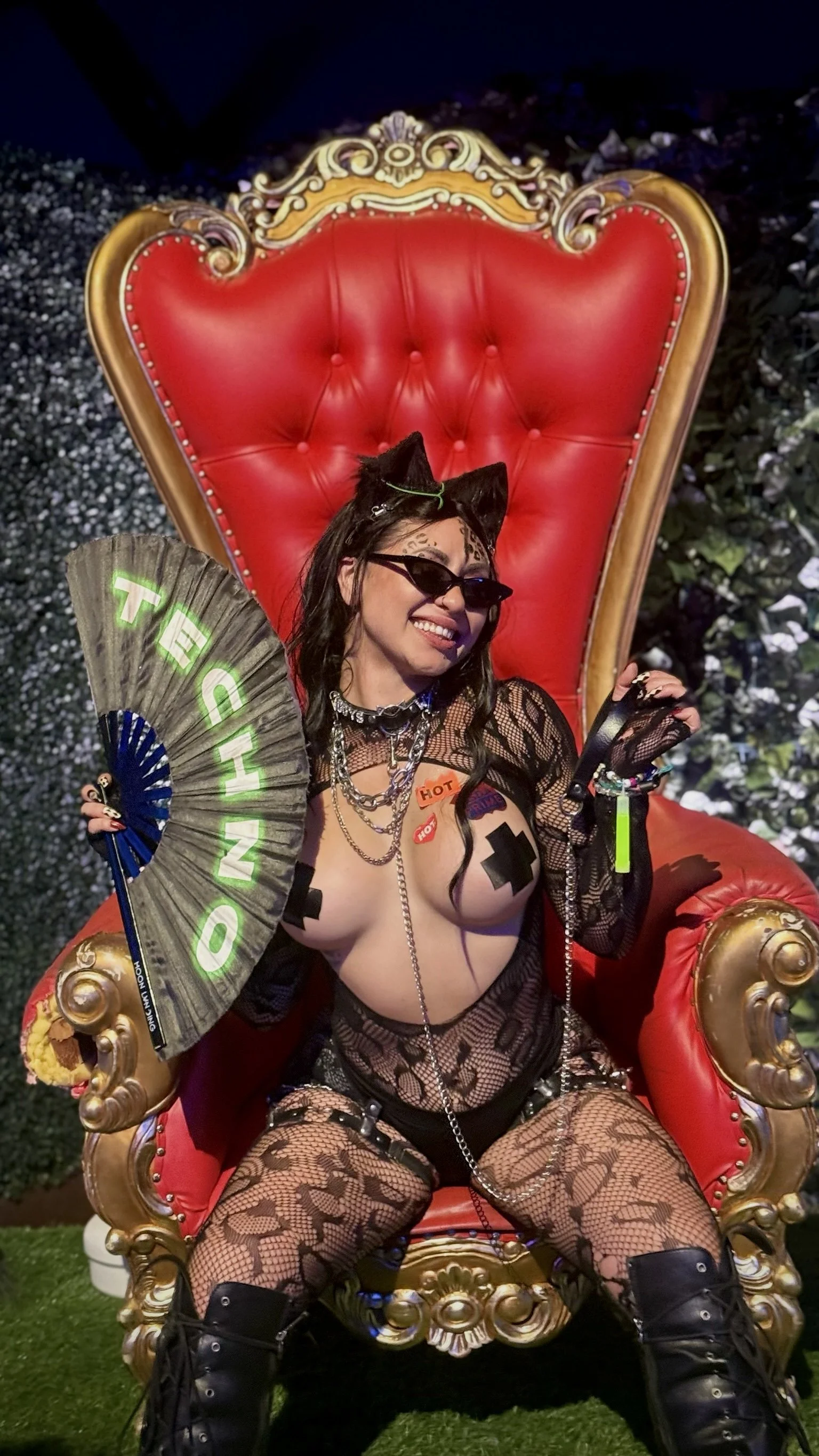 A woman dressed in provocative black fishnet clothing, with cat ears, sunglasses, and various jewelry, sitting on a large ornate red and gold throne-like chair.