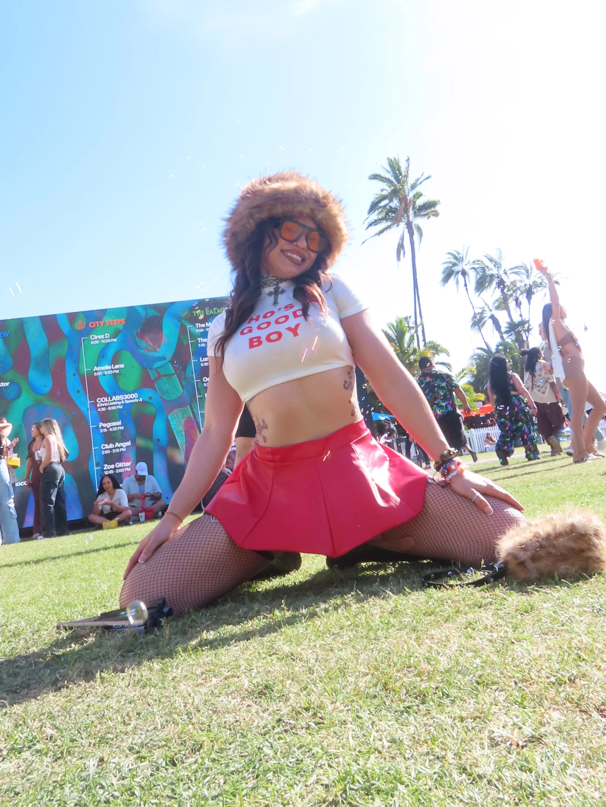 A woman outdoors on a sunny day, wearing a furry hat, sunglasses, a white crop top with red text, red skirt, and fishnet stockings, posing with her knees and hands on the ground surrounded by people and palm trees.