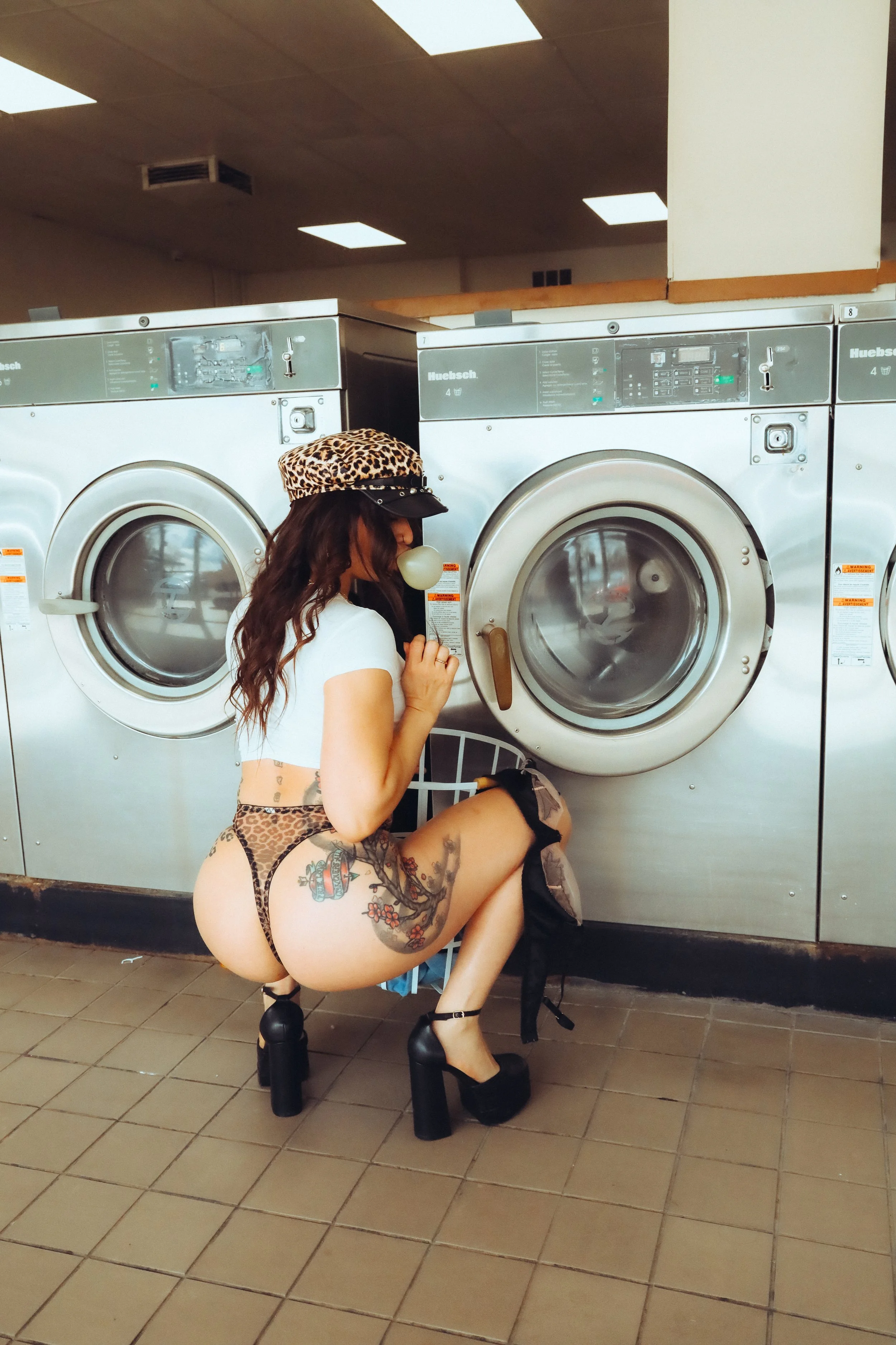 A woman with tattoos, wearing leopard print underwear, high heels, a white crop top, and a leopard print cap, crouching in front of a washing machine at a laundromat, holding and blowing bubble gum.