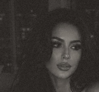 A woman with dark, curly hair, dark eye makeup, and lipstick, looking to the side in a dimly lit setting.