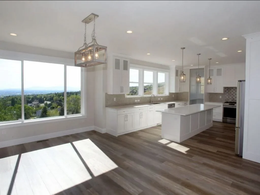 Bright, modern kitchen with large windows, white cabinetry, island, pendant lights, and a view of greenery and a distant horizon.