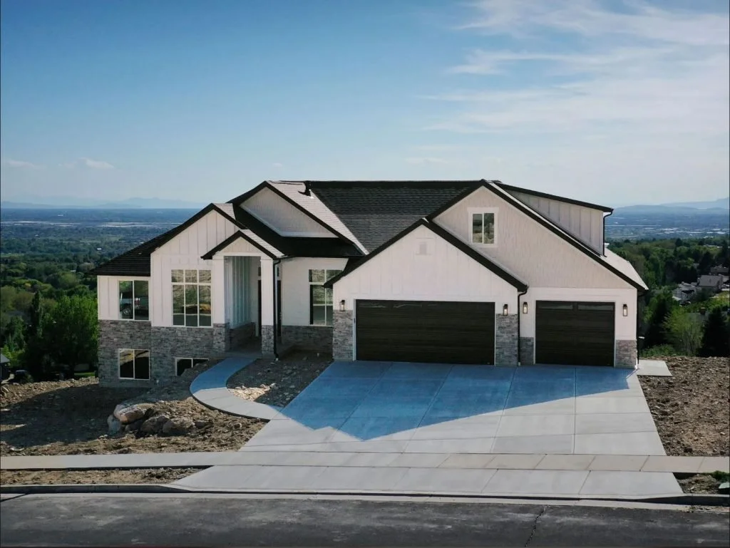 Newly built two-story house with white exterior, black roof, double garage, and large front windows, situated on a sloped lot with a concrete driveway and pathway, with a scenic view of green landscape and mountains in the background.