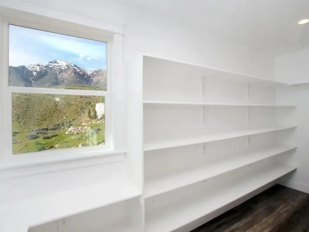 Empty white built-in shelves in a room with a window showing a mountain landscape with snow on top and grassy hills.