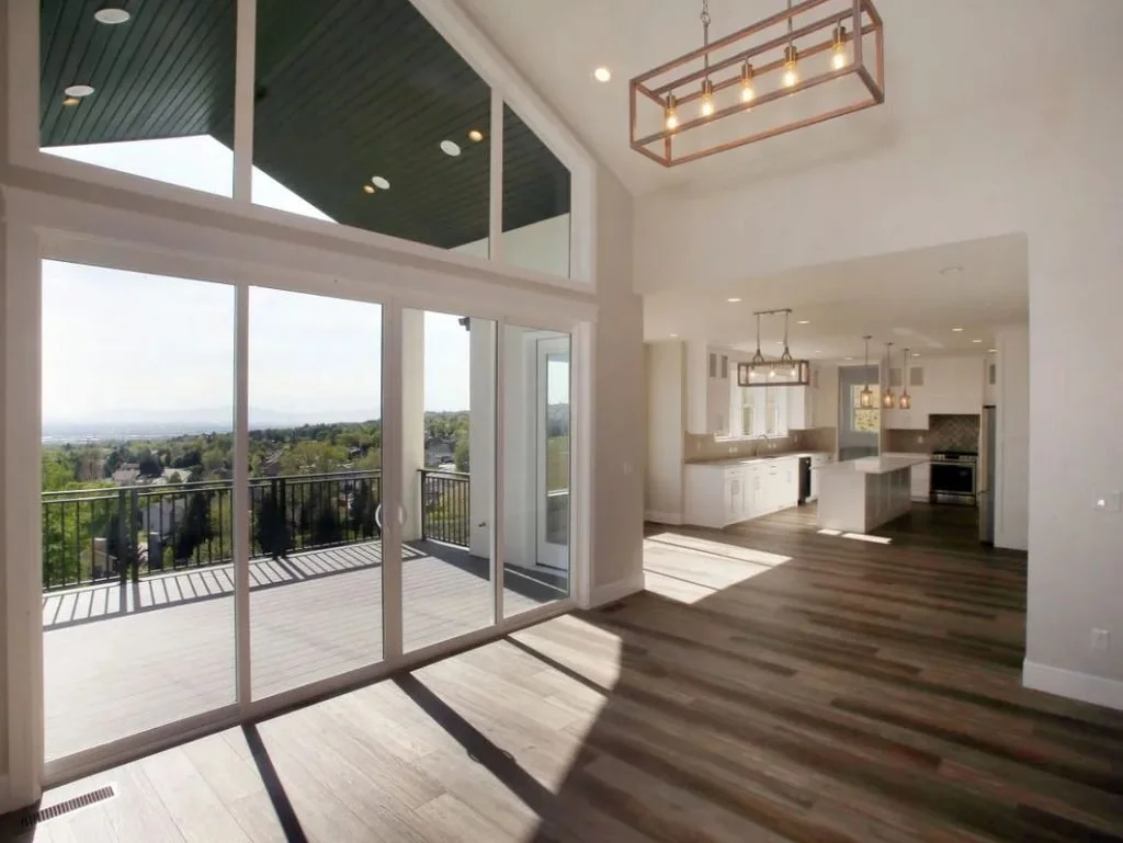 Bright, modern open-concept living room and kitchen with large glass sliding doors leading to a balcony, white cabinets, pendant lighting, and a view of distant landscape.