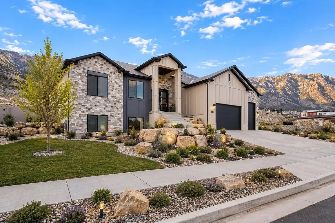 Modern house with stone and siding exterior, front yard with grass, rocks, and small plants, mountain backdrop, clear blue sky with a few clouds.