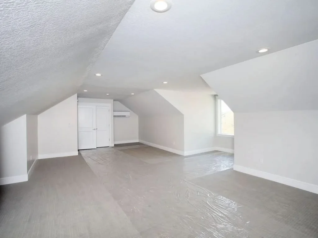 Empty attic room with sloped ceiling, white walls, a window, and recessed lighting, with a plastic sheet covering the floor.