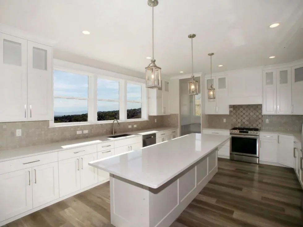 Modern spacious kitchen with white cabinets, a large white island, pendant lighting, and a view through large windows.