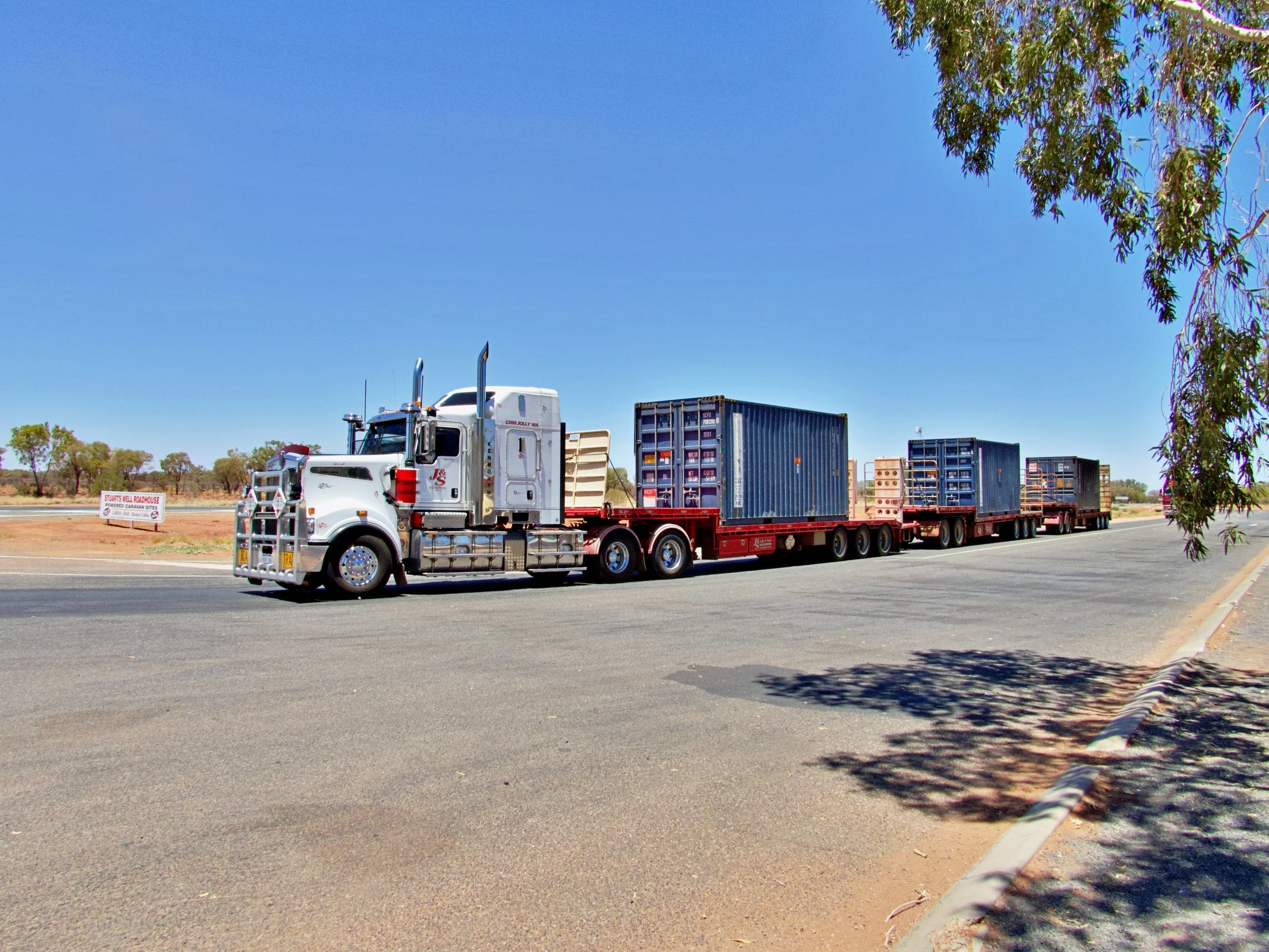 A semi-truck with a white cab and red trailer driving on a highway under a clear blue sky, with trees and a sign in the background.