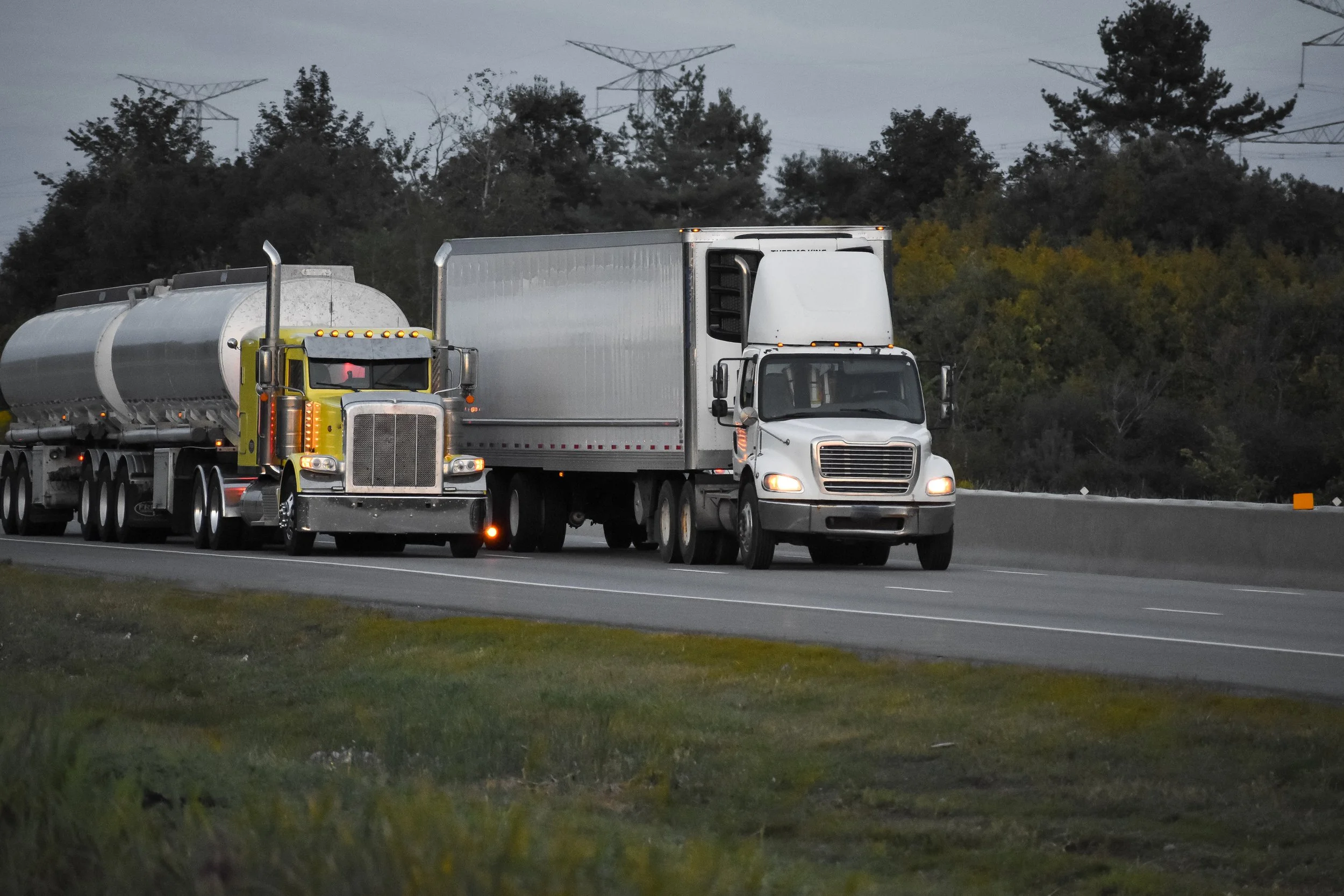 Several semi-trucks driving on a highway surrounded by trees and power lines under a cloudy sky.