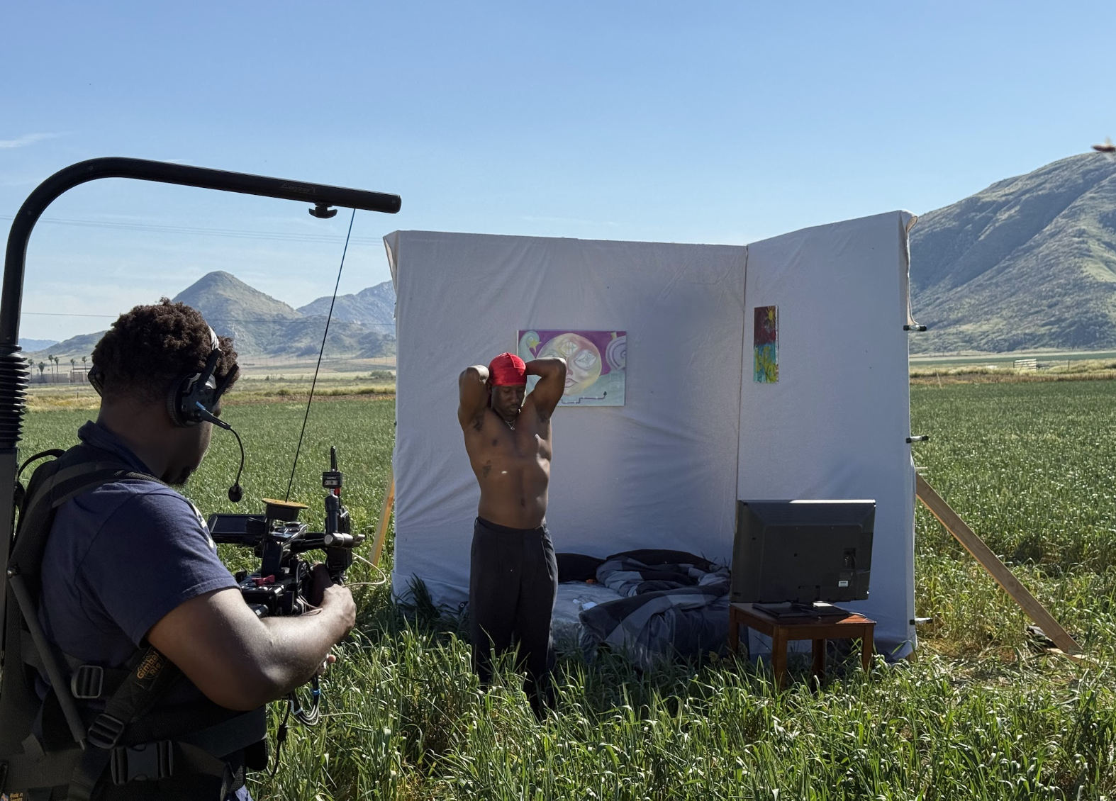 A man with red cap shirtless and standing in front of a white tarp with artwork, during outdoor photoshoot in field with mountains in background, photographer with camera on the left.