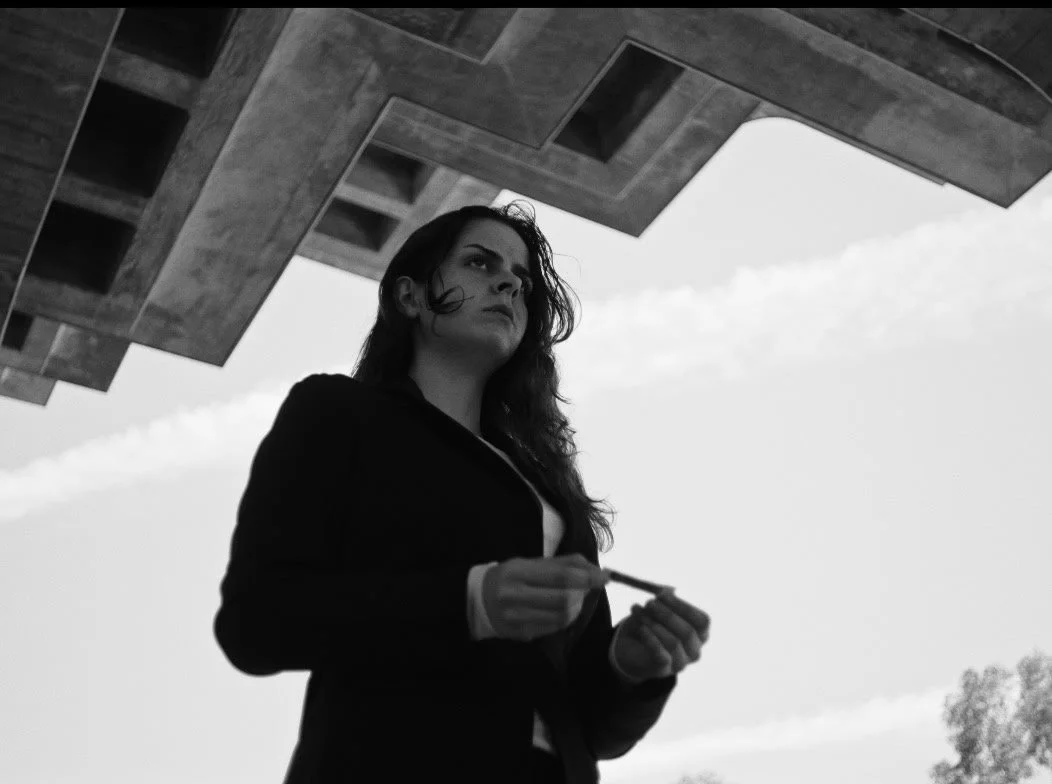 A woman with dark hair stands outdoors beneath a modern concrete structure, looking thoughtfully into the distance, holding a cigarette.