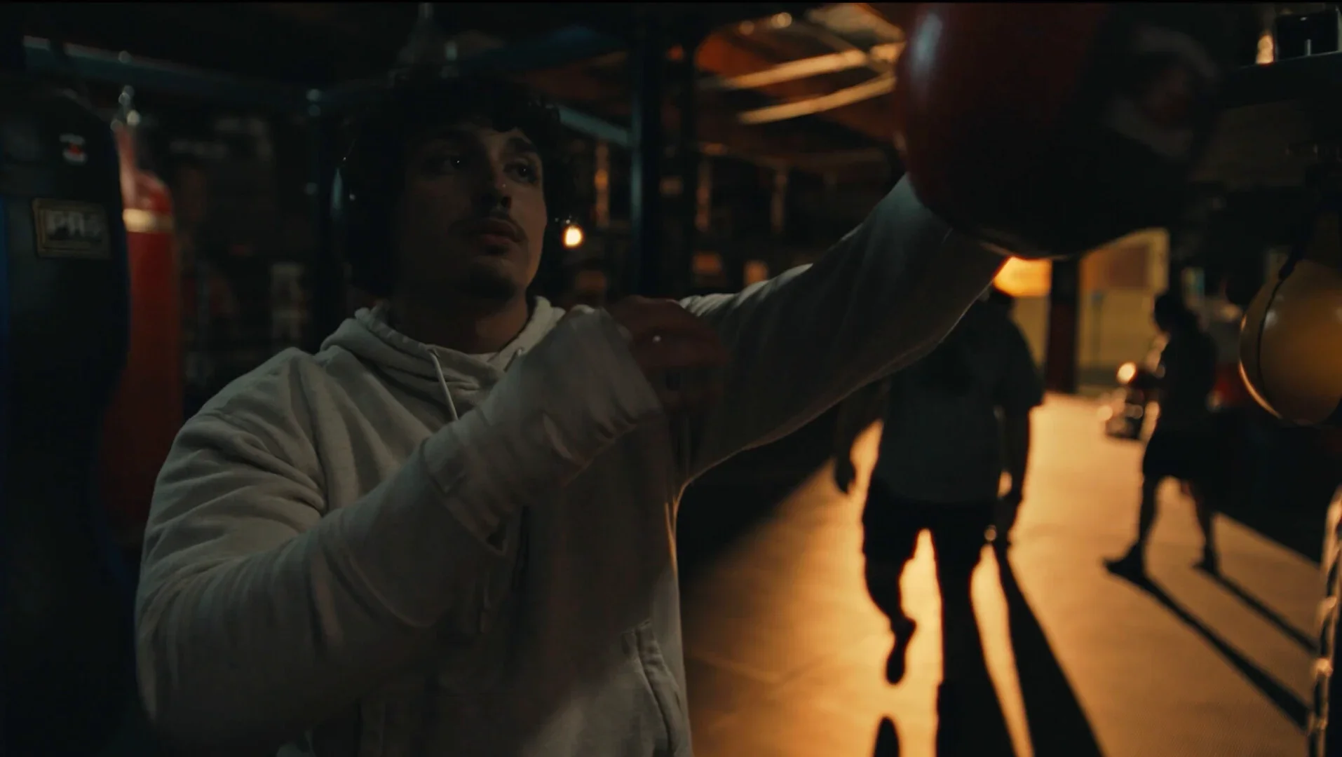 A young man wearing a hoodie and headphones adjusting his helmet in a dimly lit garage or workshop, with other people in the background.
