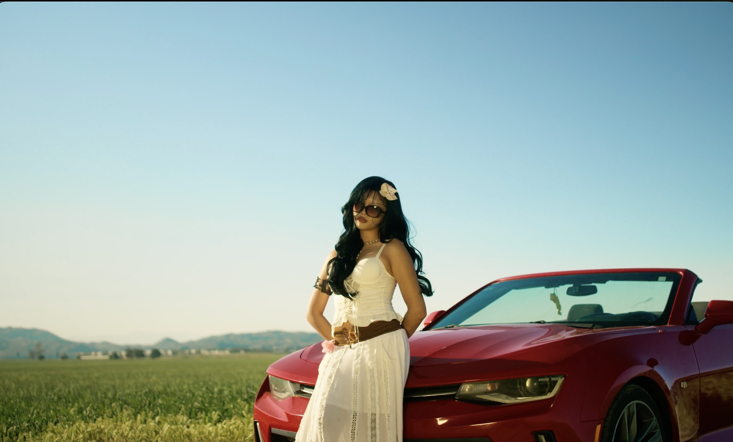 A woman in sunglasses and a white dress stands beside a red convertible car in a grassy field with mountains in the distance under a clear blue sky.