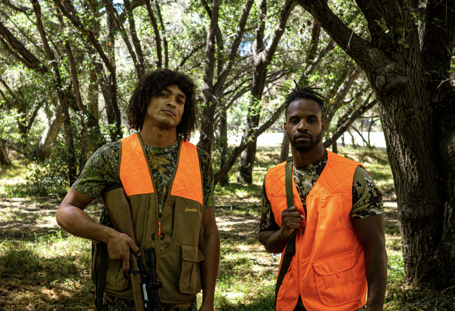 Two men wearing camouflage shirts and orange safety vests standing in a wooded area. One holds a rifle, and the other has a backpack strap.