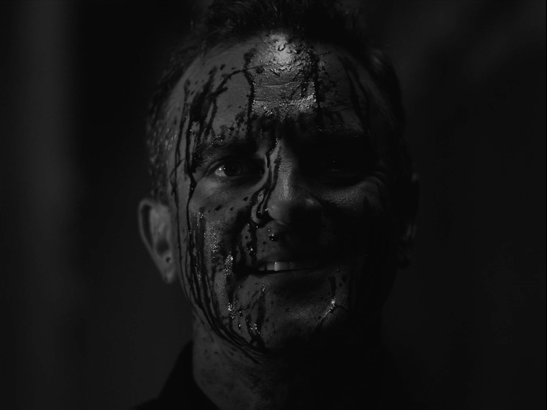 Close-up of a person’s face partially covered in black liquid, with an intense expression, wet hair, and dark background.
