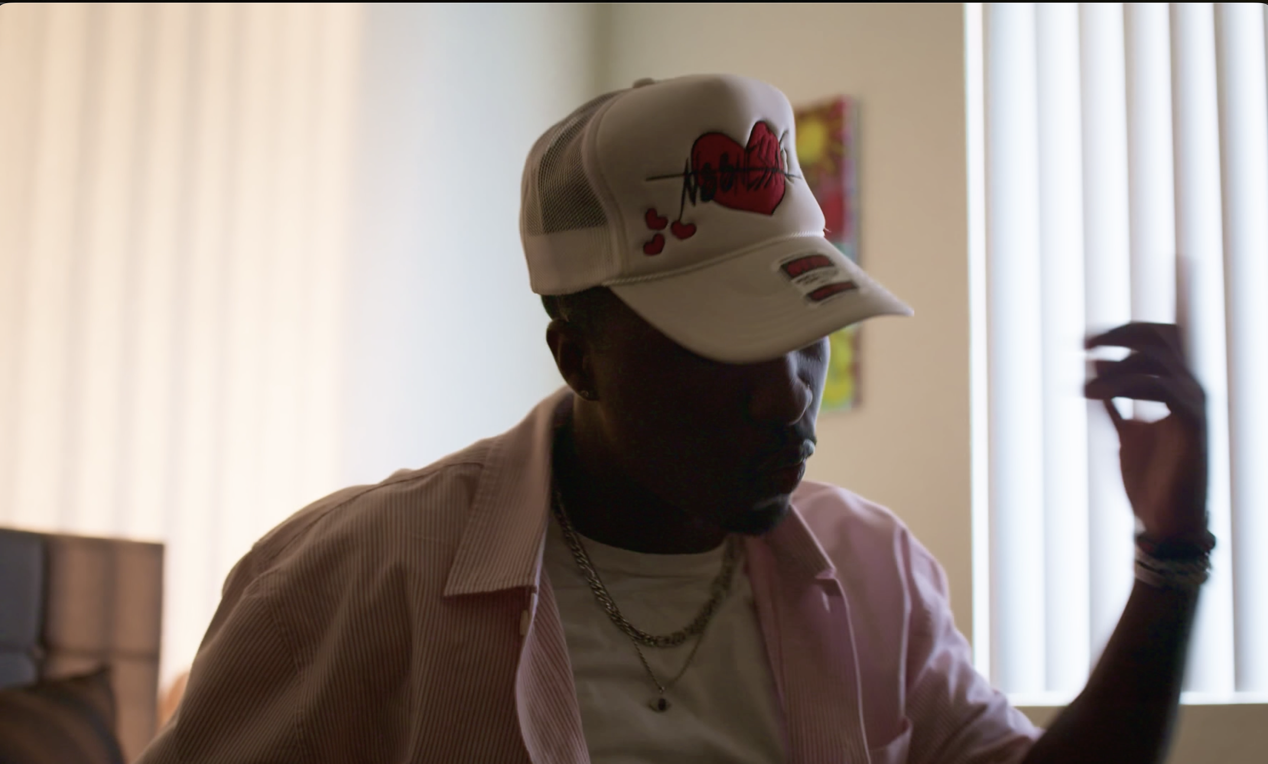 Person wearing a white cap with a red heart and black graffiti style design, inside a room with vertical blinds, facing downward, with sunglasses on, wearing a light pink shirt and a chain necklace.
