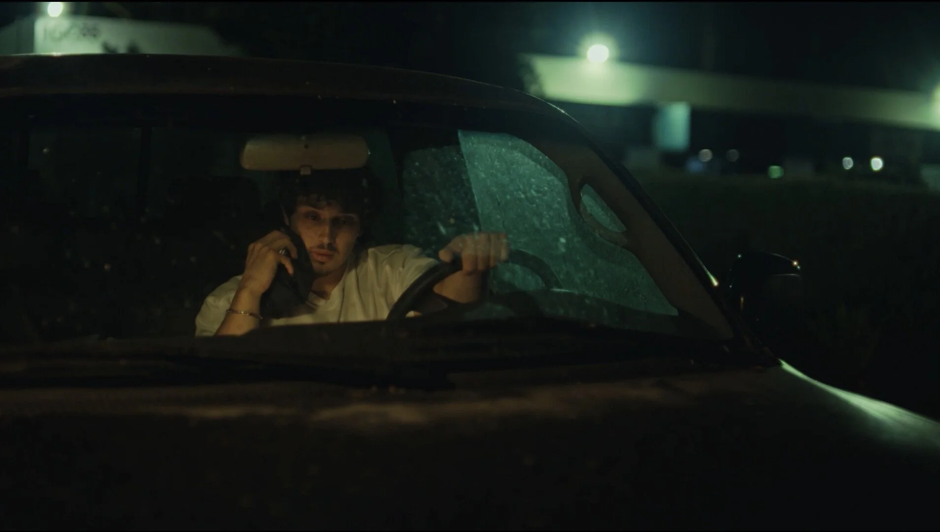 A man sitting in a car at night, speaking on a cell phone, with rain on the windshield and a bridge or overpass in the background.