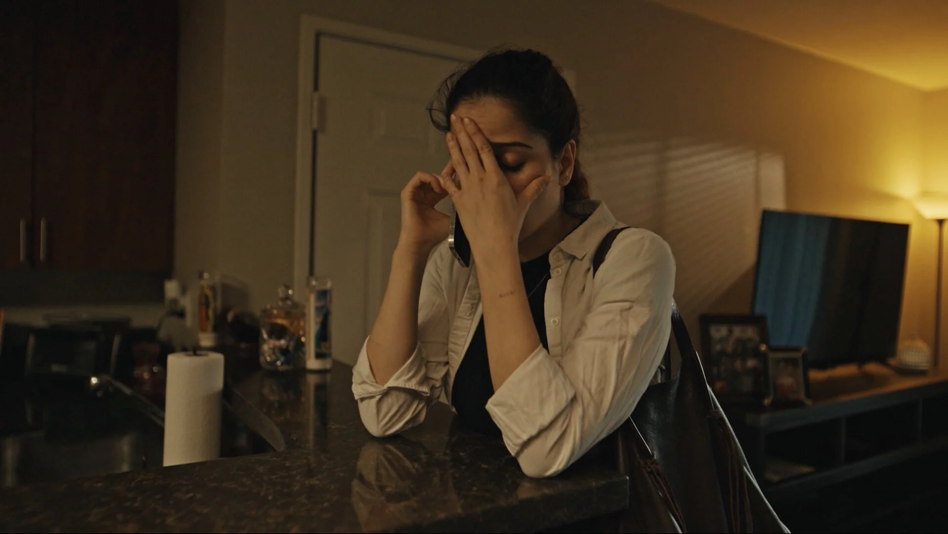 A woman sitting at a kitchen counter, holding her forehead with one hand and a phone to her ear with the other, appearing distressed.