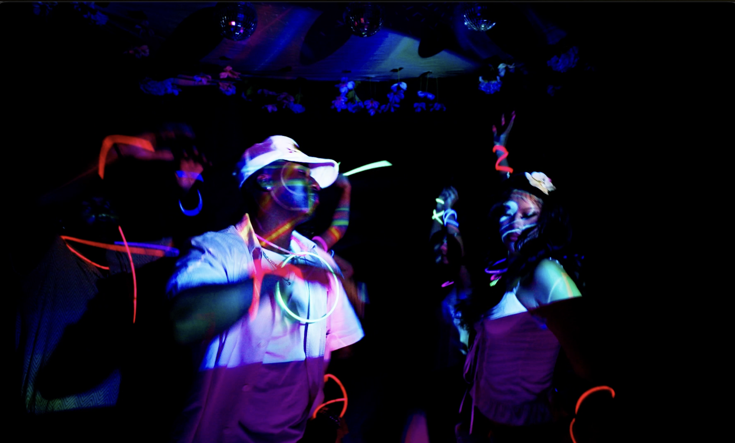 People dancing at a party under blacklights with glow sticks and neon accessories, with disco balls hanging from the ceiling.