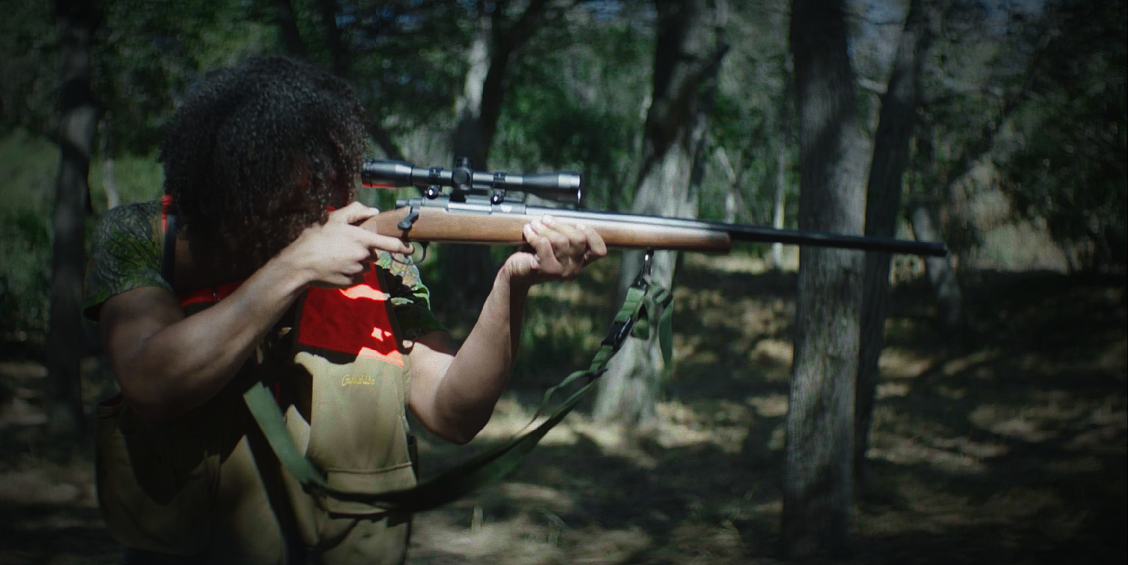 A person aiming a rifle with a scope in a wooded outdoor area during daytime.