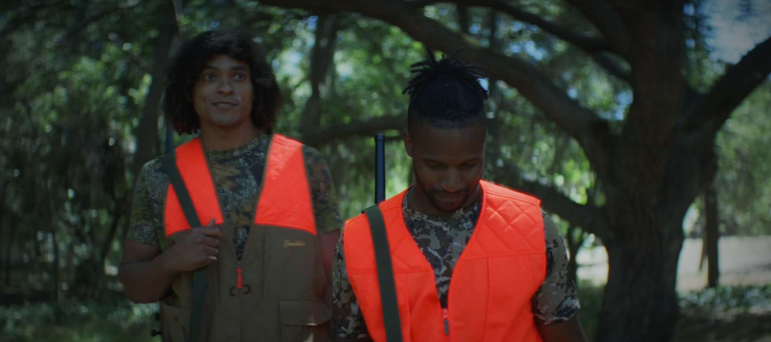 Two men walking outdoors in a wooded area, wearing orange safety vests and camouflage shirts, with one smiling and the other looking down.