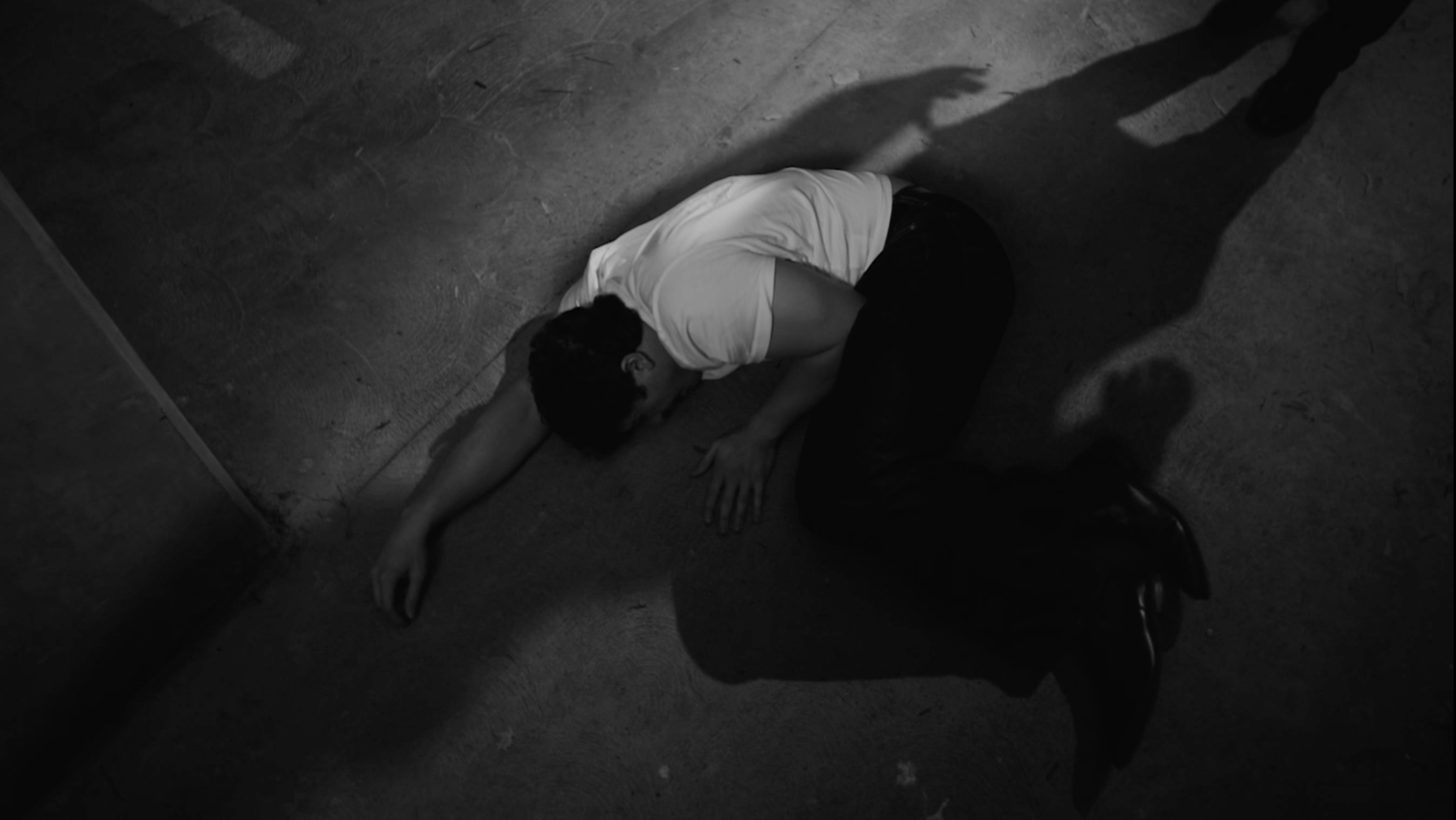 A person lies on the ground with their arm extended, their head turned to the side, and wearing a white shirt and dark pants. The scene is dimly lit with shadows cast on the floor.