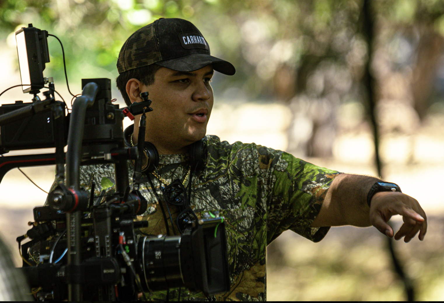 A man in camouflage shirt and baseball cap operating a camera on a tripod outdoors, with trees in the background.