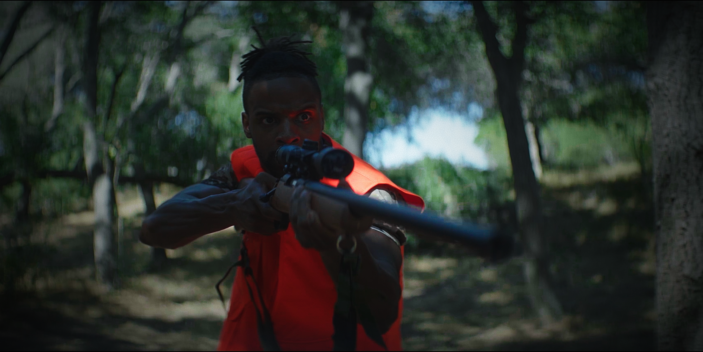A person aiming a rifle in a forest, wearing a red jacket and looking through a scope.
