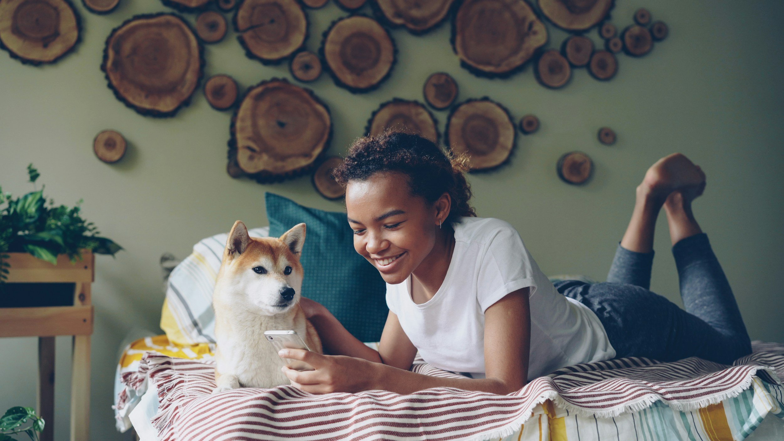 A young woman and a dog lying on a bed, smiling at a smartphone, with a wall decorated with wooden log slices in the background.