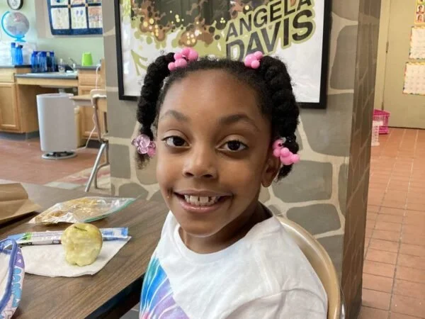A young girl with braided hair decorated with pink beads, smiling at the camera, sitting at a table with a cookie and snacks in a classroom.