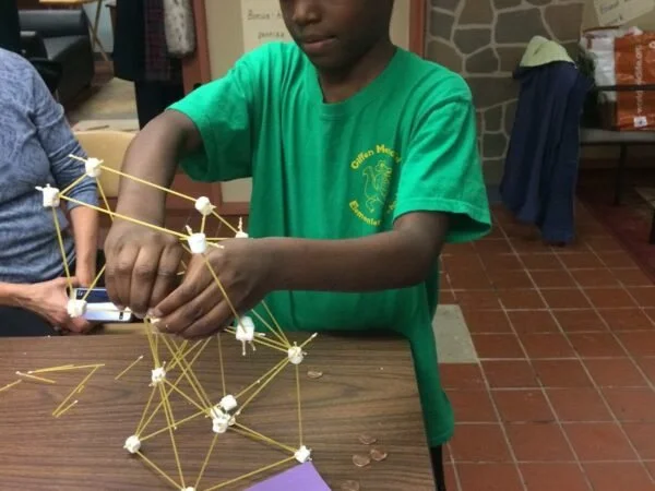 A boy in a green t-shirt building a structure with marshmallows and spaghetti sticks on a wooden table.