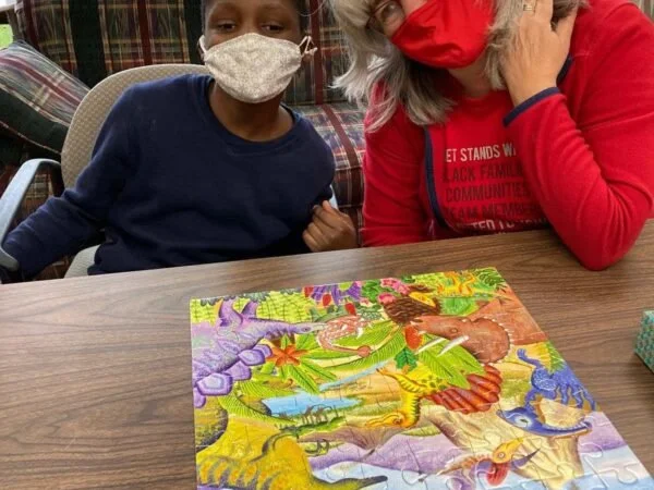 A young girl and an older woman wearing face masks sitting at a table with a colorful 500-piece jigsaw puzzle spread out in front of them.