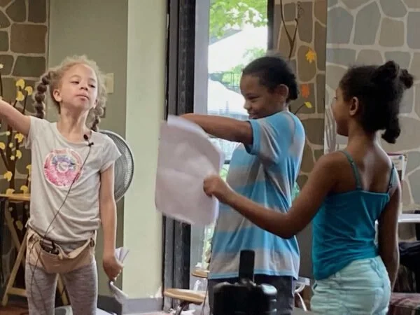 Three children standing indoors, with two girls and one boy, engaging in a playful activity. The girl on the left has curly hair, wearing a light-colored shirt with a design, and is speaking or singing. The boy in the middle is smiling, holding a pie